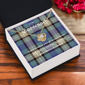 Pode incluir: Um colar dourado com um pendente de n&oacute;, apresentado numa caixa de presente branca aberta com um padr&atilde;o xadrez azul, verde e cinzento. A caixa apresenta o texto "To My Sassenach" e uma cita&ccedil;&atilde;o sobre o amor.
