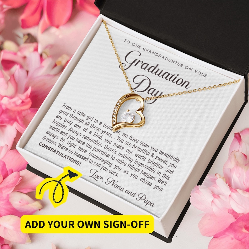 Peut inclure: Un collier pendentif en forme de c&oelig;ur dor&eacute; avec une pierre pr&eacute;cieuse transparente, pr&eacute;sent&eacute; dans un coffret cadeau blanc. La bo&icirc;te contient un message pour une petite-fille pour son jour de remise de dipl&ocirc;me. Fleurs roses en arri&egrave;re-plan.