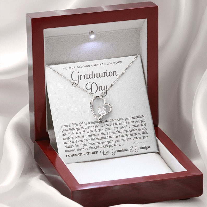 Peut inclure: Un collier en argent avec un pendentif en forme de c&oelig;ur orn&eacute; d'une pierre pr&eacute;cieuse transparente est pr&eacute;sent&eacute; dans un coffret &agrave; bijoux en bois rouge ouvert. Le coffret contient une carte blanche avec le texte "To our Granddaughter on your Graduation Day."