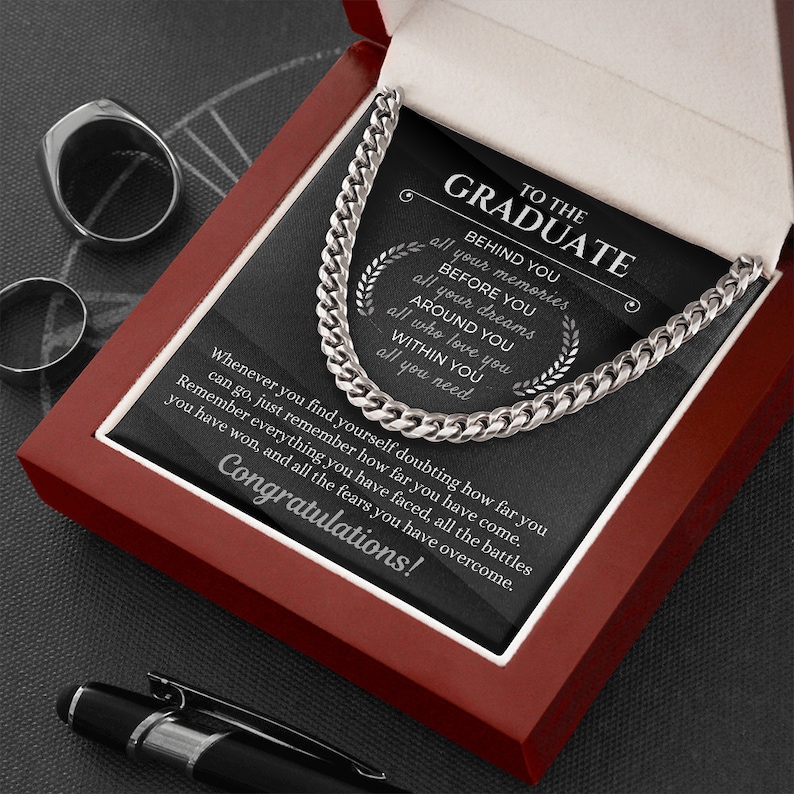 Pu&ograve; includere: Una collana a catena d'argento in una scatola di velluto rosso con una placca nera su cui &egrave; scritto "To the Graduate" e un messaggio motivazionale sul superamento delle sfide e il raggiungimento degli obiettivi.