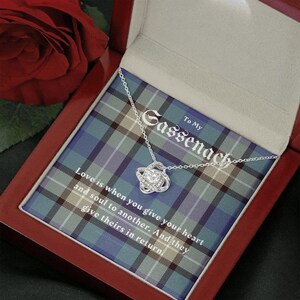 Pode incluir: Um colar prateado com um pendente de n&oacute;, apresentado numa caixa de presente vermelha. O interior da caixa apresenta um padr&atilde;o xadrez azul e verde com o texto "To My Sassenach". O texto "Love is when you give your heart and soul to another. And they give theirs in return" tamb&eacute;m &eacute; vis&iacute;vel.