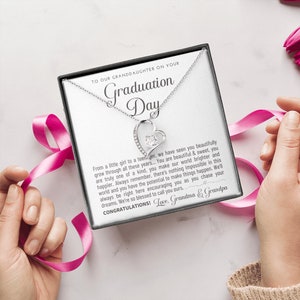 Peut inclure: Un collier pendentif argent&eacute; en forme de c&oelig;ur avec une pierre pr&eacute;cieuse transparente, pr&eacute;sent&eacute; dans un coffret cadeau. Le coffret porte l'inscription "TO OUR GRANDDAUGHTER ON YOUR Graduation Day" et un message. Un ruban rose entoure le coffret.