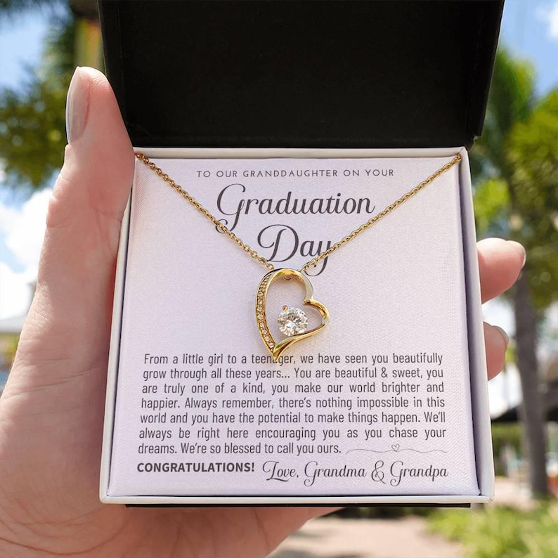 Peut inclure: Un collier pendentif en forme de c&oelig;ur dor&eacute; avec une pierre pr&eacute;cieuse transparente, pr&eacute;sent&eacute; dans un coffret cadeau. Le coffret contient un message "TO OUR GRANDDAUGHTER ON YOUR Graduation Day". Le message est de Grand-m&egrave;re et Grand-p&egrave;re.
