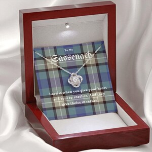 Pode incluir: Um colar de prata com um pendente de n&oacute;, apresentado numa caixa de presente de madeira. A caixa cont&eacute;m um cart&atilde;o com o texto "To My Sassenach" e uma cita&ccedil;&atilde;o sobre o amor. O fundo apresenta um padr&atilde;o xadrez azul, verde e branco.