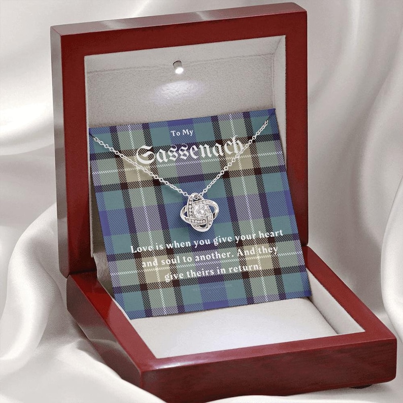 Pode incluir: Um colar de prata com um pendente de n&oacute;, apresentado numa caixa de presente de madeira. A caixa cont&eacute;m um cart&atilde;o com o texto "To My Sassenach" e uma cita&ccedil;&atilde;o sobre o amor. O fundo apresenta um padr&atilde;o xadrez azul, verde e branco.