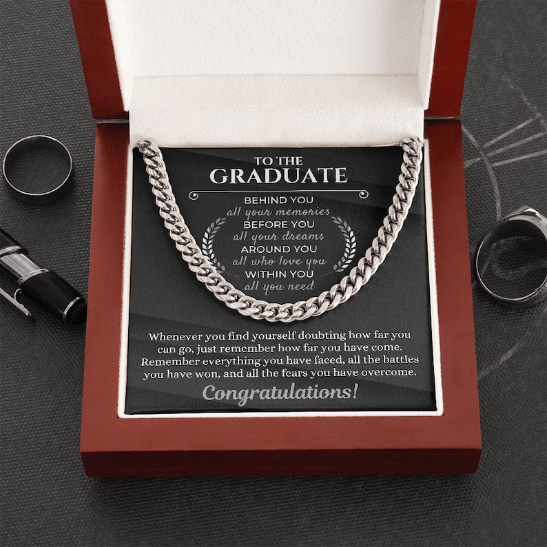 Pu&ograve; includere: Una collana con catena d'argento in una scatola di velluto rosso con sfondo nero. La scatola contiene un messaggio per un laureato che recita "Al laureato" e include parole di ispirazione su ricordi, sogni e amore.