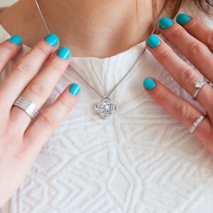 Pode incluir: Um colar de prata com um pingente de n&oacute;, incrustado com pequenas pedras preciosas transparentes. O colar &eacute; exibido em um top branco texturizado. M&atilde;os com esmalte de unhas turquesa s&atilde;o vis&iacute;veis, adornadas com an&eacute;is de prata.