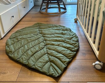May include: A large green leaf-shaped play mat made of quilted fabric. The mat is on a wooden floor.