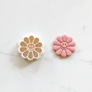 May include: A white plastic cookie cutter and a pink cookie in the shape of a daisy with a smiley face in the center.