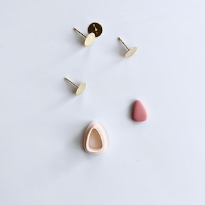 Puede incluir: Un conjunto de suministros para pendientes que incluye tres postes de pendientes dorados con respaldos planos, una pieza de arcilla de forma triangular rosa y un molde de plástico blanco para hacer pendientes.