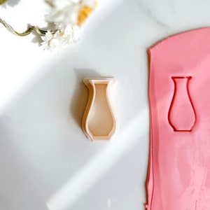 May include: A white plastic cookie cutter shaped like a vase. The cutter is on a white surface next to a pink fondant sheet with a vase shape cut out.