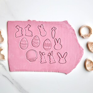 May include: A set of 15 plastic cookie cutters in various Easter shapes, including bunnies, eggs, baskets, and carrots. The cutters are arranged on a pink fondant surface and a white marble countertop.