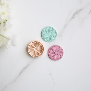 May include: Three silicone cookie cutters shaped like a slice of citrus fruit. The cutters are in different colors: light blue, light pink, and beige. The beige cutter is open, showing the inside of the cutter.