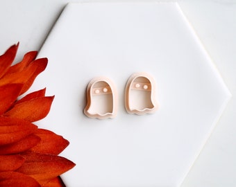 May include: Two light beige ghost-shaped cookie cutters. Each cutter has a rounded top with two small circular eye holes and a wavy bottom edge. The cutters are on a white hexagonal surface.
