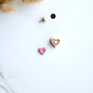 May include: Two pink heart-shaped earrings with a smaller heart cutout in the center. The earrings are on a white surface.