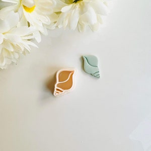 May include: Two plastic cookie cutters, one shaped like a seashell in light brown and the other shaped like a seashell in light blue.