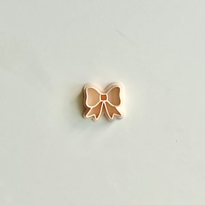 Puede incluir: Un cortador de galletas de plástico con forma de lazo. El cortador es de color melocotón claro.