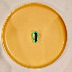 May include: A green plastic cookie cutter shaped like a shield on a tan speckled plate.