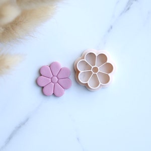 Peut inclure: Un emporte-pièce en forme de fleur rose clair à côté d'une fleur en fondant assortie. L'emporte-pièce est en plastique beige clair. La fleur en fondant a un centre et un design de pétales détaillés. Le fond est une surface en marbre blanc.