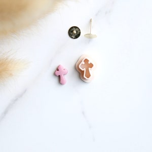 May include: Two pink cross-shaped charms and a pair of earring findings on a white marble surface.