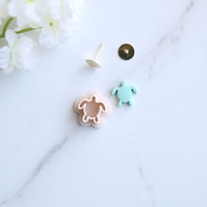 May include: A set of two sea turtle earrings, one is a light blue plastic turtle and the other is a pink plastic mold of a turtle. The set also includes two gold earring posts and two gold earring backs.