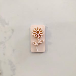 Puede incluir: Un sello de plástico blanco con un diseño de flor. La flor tiene un centro circular y pétalos que se extienden hacia afuera. La flor está en un tallo con dos hojas.
