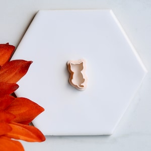 May include: A light beige cat-shaped cookie cutter sits on a white hexagonal tile. The cutter is a silhouette of a sitting cat. An orange flower is in the lower left corner of the image.