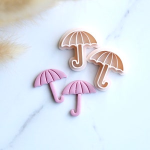 Puede incluir: Cuatro cortadores de galletas con forma de paraguas. Dos son de color rosa y dos son de color beige claro.