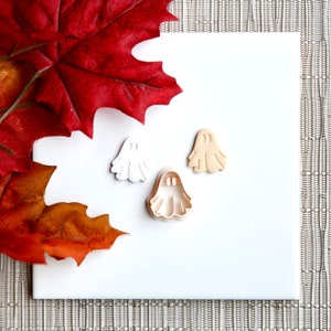 May include: Three ghost-shaped cookie cutters in white and tan, with a red and orange leaf arrangement. The cutters are on a white surface, with a textured background.