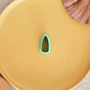 May include: A teal-colored teardrop-shaped cookie cutter sits on a yellow plate. The plate is on a textured, neutral-colored surface. A decorative dried flower element is in the upper right corner. The cookie cutter is a simple, open design.