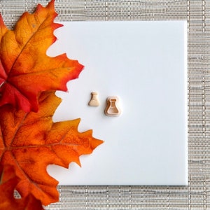 May include: Two small, beige plastic cookie cutters shaped like beakers on a white surface with a fall-colored leaf in the background.