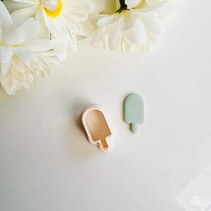 May include: Two popsicle-shaped cookie cutters, one light peach and one pale green, sit on a white surface. White flowers are in the background. The cookie cutters are for making treats.