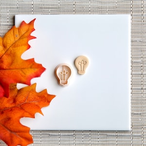 May include: Two light bulb shaped cookie cutters, one larger than the other, on a white surface with a fall colored leaf in the background.