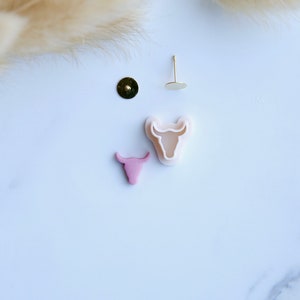 May include: A set of two white plastic bull head cookie cutters, one with a pink bull head inside. The cutters are on a white surface with a gold earring post and a gold earring back.