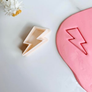 May include: A white plastic lightning bolt cookie cutter with a pink play dough impression of the same shape.