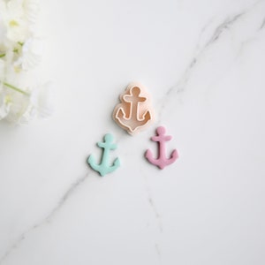 May include: Three anchor-shaped cookie cutters, one is white, one is light blue, and one is pink. The cutters are on a white marble surface.