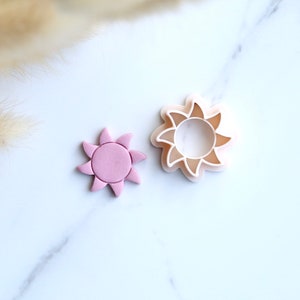 Puede incluir: Dos cortadores de galletas con forma de sol. El cortador más grande es blanco y el más pequeño es rosa.