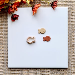 May include: A white surface with a cookie cutter shaped like a fish, a light brown clay fish, and a dark brown clay fish.