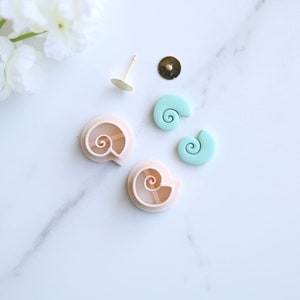 Op de afbeelding: Twee lichtroze schelpvormige oorknopvormen van polymer clay en twee lichtblauwe schelpen van polymer clay. De schelpen zijn klaar om als oorknoppen te worden gebruikt. De vormen zijn voor het maken van oorknoppen.