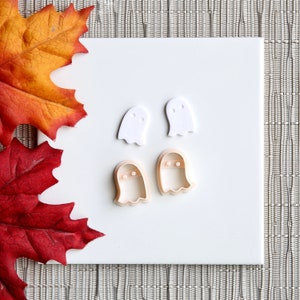 May include: Four white and peach-colored ghost-shaped cookie cutters on a white surface. The cookie cutters are for making Halloween treats.