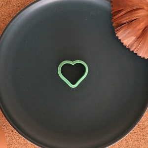 Puede incluir: Un cortador de galletas en forma de corazón verde sobre un plato negro. El plato está sobre una superficie de corcho con una flor marrón.