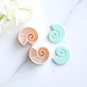 May include: Two plastic cookie cutters shaped like seashells with a spiral design. Two light blue fondant seashells are also shown.