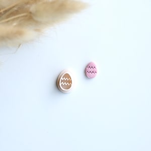 May include: Two Easter-themed items on a white surface. One is a light pink egg-shaped cookie cutter with a wavy design inside. The other is a small, light pink egg-shaped decoration with a wavy pattern. Dried plants are in the background.