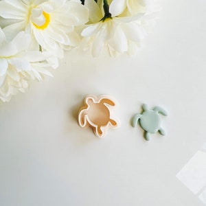 May include: Two plastic cookie cutters in the shape of sea turtles. One is light green and the other is light pink.