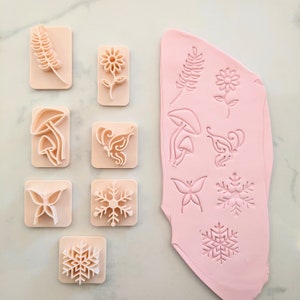 K&ouml;nnte beinhalten: Ein Set mit 10 Silikonformen zum Backen und Basteln. Die Formen zeigen verschiedene Designs, darunter Schmetterlinge, Pilze, Schneeflocken und Bl&auml;tter. Die Formen sind rosa und haben eine glatte, gl&auml;nzende Oberfl&auml;che.