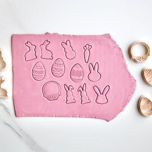 May include: A set of 15 plastic cookie cutters in various Easter shapes, including bunnies, eggs, baskets, and carrots. The cutters are arranged on a pink fondant surface and a white marble countertop.