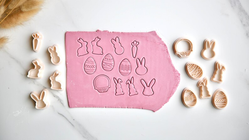 May include: A set of 15 plastic cookie cutters in various Easter shapes, including bunnies, eggs, baskets, and carrots. The cutters are arranged on a pink fondant surface and a white marble countertop.