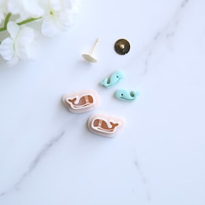 May include: A set of four whale-shaped earrings, two are light blue and two are a pale pink colour. The earrings are on a white surface.