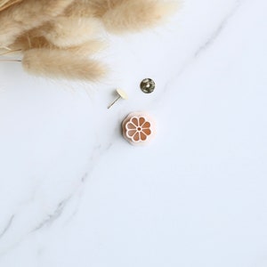 May include: A collection of jewelry components, including a small, light pink flower-shaped charm, a gold-colored earring post, and a small, round, textured gold-colored stud. Dried, beige-colored decorative grass is in the upper left corner. The items are arranged on a white marble surface.