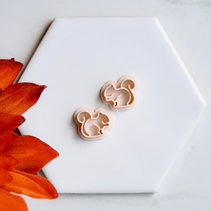 May include: Two light pink plastic squirrel cookie cutters on a white hexagonal surface.